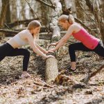 Side view of young female athletes in activewear stretching legs together using fallen tree during fitness workout in spring forest adhering to healthy lifestyle